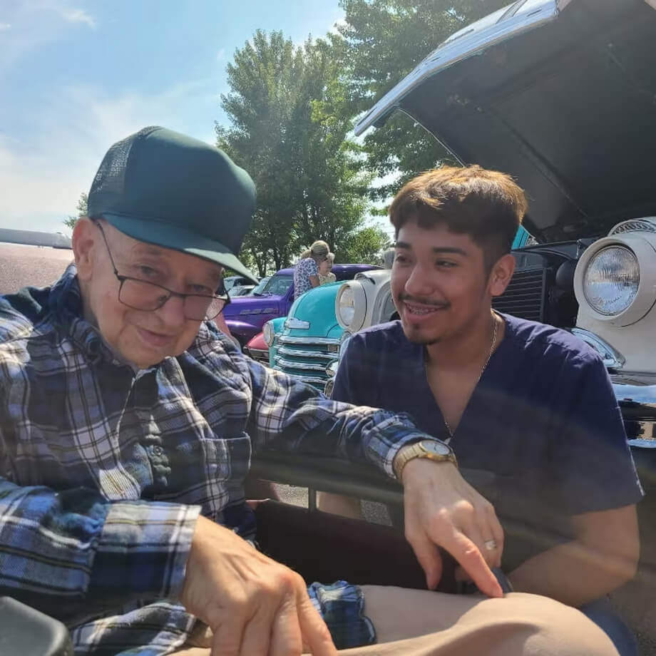 Caregiver spending time with an older man at an outdoor classic car event.